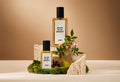 The image shows a soft, beige background. In the centre of focus are two bottles of two different sizes of perfume sitting on top of moss and rock with moss, blackcurrant and litsea cubeba flowers.